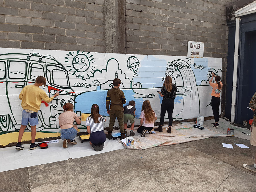 A team of school-aged children are painting a large mural on a brick wall. The mural depicts a red Kombi van on a beach with an ocean view.