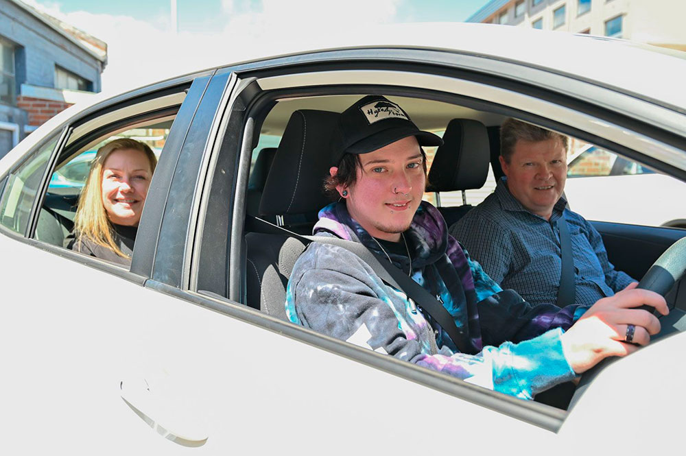 A group of adults and a young person posing for a photo inside a car. They are smiling and wearing casual clothes. The image is promoting a program that helps young people learn how to drive safely.