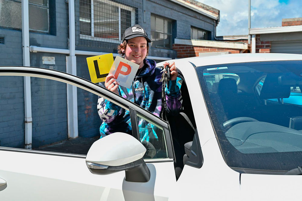 A young individual stands next to the driver's side door, smiling. They hold keys in one hand and an L plate and P plate in the other.